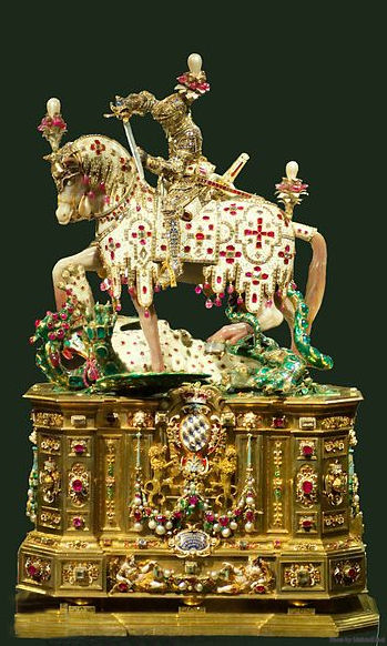 Duke Wilhelm’s son, Maximilian I, had the original ebony base replaced with the present sumptuous pedestal. The entire statuette consists of 2,291 diamonds, 406 rubies, and 209 pearls. At the base, the inscription in gold letters reads: “Maximilian, Count Palatine on the Rhine, Duke of the two Bavarias, Elector of the Holy Roman Empire, dedicated this to the great martyr St. George, patron and protector of his house and family.” Well concealed by this lavish decoration is a tiny drawer containing a reliquary of St. George jeweled as elaborately as all the rest. This statue is housed in the Residenz Museum in Munich, Germany. The tiny statuette is barely 20″ high from its base to the pearl on the knights helmet.