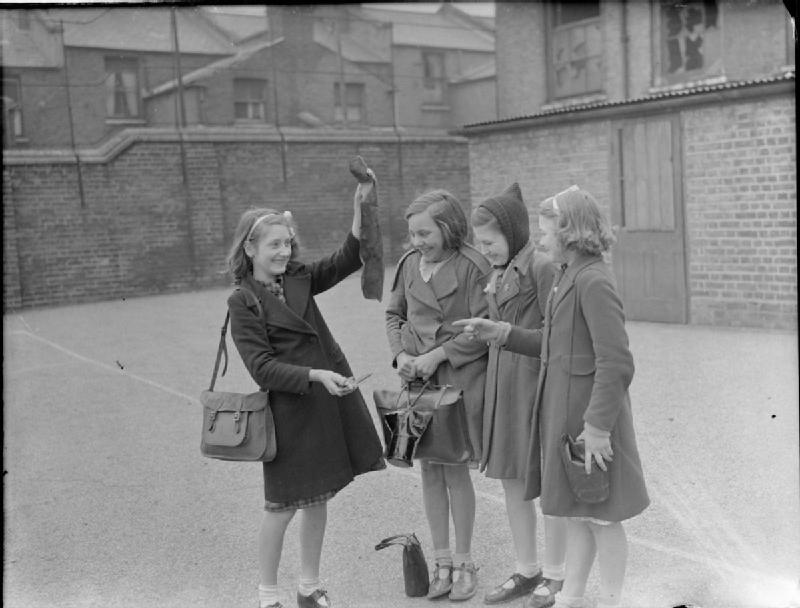A group of four smiling girls examine some pieces of metal in the playground of Calvert Road School in South East London. This debris appears to be fragments of a bomb, or of a metal structure damaged or destroyed by an air raid. One girl (second from the right) is wearing a Girl Guide badge, and all are carrying gas masks.
